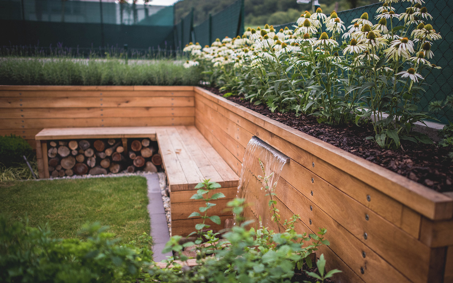 Vyvýšený záhon spojený s lavičkou, vysadený okrasnými rastlinami, ktoré zakrývajú nevzhľadný plot suseda.